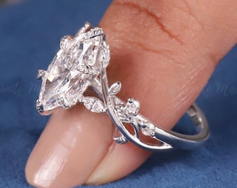 May include: A silver-toned ring featuring a large marquise-cut clear gemstone as the centerpiece. The band is designed with intricate metalwork resembling delicate vines and leaves, accented with small, sparkling clear stones along the sides.
