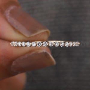 May include: A delicate gold band ring set with small, sparkling clear gemstones. The ring is held against a gray background, showcasing its fine details and elegant design. This ring is a piece of fine jewelry.