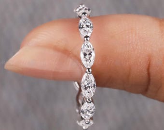 Puede incluir: Un anillo de plata con una serie de piedras preciosas de talla marquesa. El anillo se sostiene entre dos dedos sobre un fondo gris. Las piedras preciosas son claras y brillantes. Es una joya.