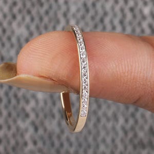 Könnte beinhalten: Ein goldfarbener Ring mit einem Band, das mit kleinen, klaren Edelsteinen besetzt ist. Der Ring wird von einem Finger vor einem strukturierten grauen Hintergrund gehalten. Der Ring ist ein zarter Schmuck.