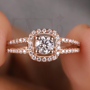 May include: Rose gold engagement ring with a square halo design. The ring features a central, round-cut diamond surrounded by smaller stones. The band splits into two rows of pave-set diamonds, adding sparkle and detail to the jewelry.