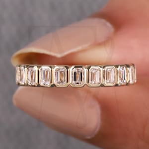 May include: A gold ring with a continuous row of emerald-cut gemstones. The ring's design showcases a series of rectangular stones set closely together, creating a sparkling, elegant band. The ring is held up against a neutral background.