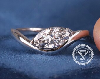 May include: A silver-toned ring featuring a single marquise-cut clear gemstone set in a smooth, curved band. The ring is positioned on a blue velvet surface and includes a small circular logo with the text IGI and a diamond graphic.