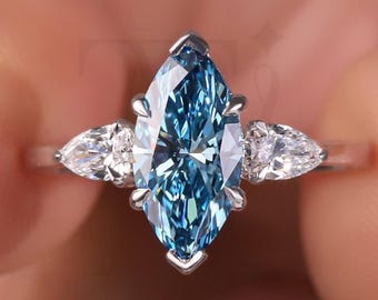 May include: A marquise-cut blue gemstone ring with two pear-shaped white gemstones on a silver band. The ring is held between fingers, showcasing the ring's design and color. The ring is a piece of jewelry.