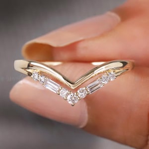 May include: A gold-colored V-shaped ring with small, clear gemstones. The ring has a delicate design, with a row of round and baguette-cut stones. The ring is held between fingers, showcasing its elegant shape and sparkle.