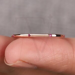 May include: A thin, rose gold ring with three small, vibrant pink gemstones set across the top. The ring is held between two fingers, showcasing its delicate design and minimalist style. The ring's polished surface reflects light, highlighting its elegant simplicity.