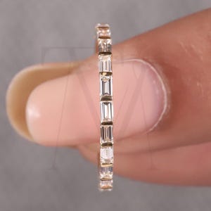 以下が含まれることがあります： 長方形の透明な宝石が連続して並んだ繊細なゴールドリング。指の間にリングが挟まれ、その繊細なディテールとエレガントなデザインが際立っています。宝石は均等に配置され、輝くバンドを形成しています。