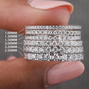 Pode incluir: Uma pilha de sete anéis de prata com pedras preciosas transparentes, cada um com uma largura diferente, variando de 1,00 mm a 2,50 mm. Os anéis são segurados entre os dedos, mostrando os vários tamanhos. Os anéis são da Moissanite.