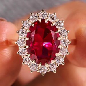 Könnte beinhalten: Ein Goldring mit einem großen, oval geschliffenen, tiefroten Edelstein in der Mitte, umgeben von einem Kranz aus kleinen, runden, klaren weißen Steinen. Die Fassung besteht aus einem polierten Gelbgoldband mit klassischem Blumendesign.