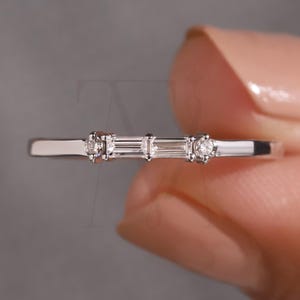 Könnte beinhalten: Ein zarter Silberring mit drei klaren Edelsteinen. Der zentrale Stein ist rechteckig, flankiert von zwei runden Steinen. Der Ring wird vor einem neutralen Hintergrund präsentiert, der sein minimalistisches Design und seine feine Verarbeitung hervorhebt.