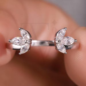 May include: A silver-toned open ring featuring two symmetrical clusters of marquise-cut clear stones. Each cluster consists of three stones arranged in a fan-like pattern at the ends of the band, creating a gap in the center of the ring design.