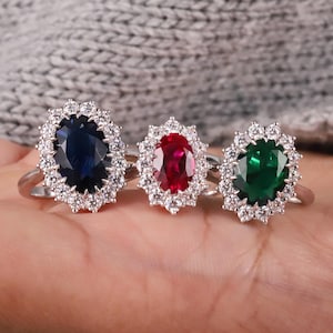 Może przedstawiać: Trzy pierścionki ze srebrnymi obrączkami, każdy z owalnym kamieniem szlachetnym w kolorze szafirowym, rubinowym i szmaragdowym, otoczonym aureolą mniejszych, przezroczystych kamieni. Pierścionki są prezentowane na dłoni.