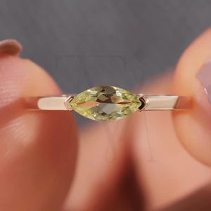 May include: A ring featuring a single marquise-cut, pale yellow gemstone set in a smooth, polished metal band. The stone is positioned horizontally, held by two prongs on either side, showcasing a faceted surface that reflects light clearly.