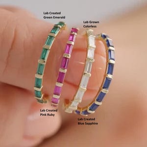 Puede incluir: Cuatro anillos de oro con piedras preciosas creadas en laboratorio: esmeralda verde, incolora, rubí rosa y zafiro azul. Cada anillo presenta una fila de piedras de corte rectangular. Los anillos están etiquetados.