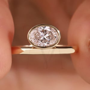 May include: A gold ring featuring a large, oval-cut diamond. The diamond is set in a bezel setting, and the band is a simple, polished gold. The ring is held between two fingers, showcasing the design and sparkle of the gemstone.