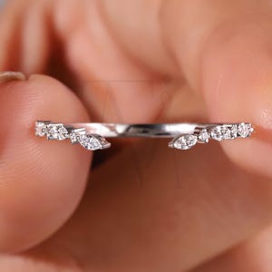 May include: A silver ring with a row of small, clear gemstones. The ring has a delicate design with a curved shape and is held between fingers. The gemstones are of varying shapes, including round and marquise cuts.