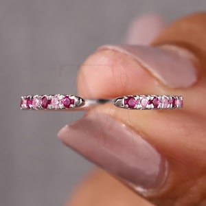 May include: A silver ring with a row of alternating pink and red gemstones. The ring is held between fingers, showcasing the gemstones' color and sparkle. The ring's design is simple, with the gemstones set closely together.