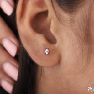 Puede incluir: Primer plano de un pequeño pendiente de botón con forma de marquesa engastado en metal plateado. El pendiente está en una oreja. Se ve una mano con esmalte de uñas rosa.