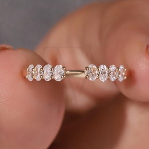 May include: A gold ring featuring a series of oval-cut diamonds. The diamonds are set closely together, creating a continuous sparkle. The ring is held between fingers, showcasing its design and craftsmanship. The ring is a piece of fine jewelry.