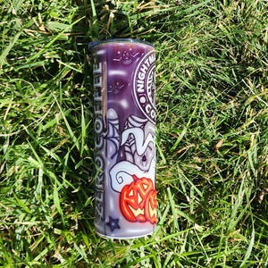 May include: A white and purple tumbler with a Halloween-themed design. The design features a ghost, a spider web, and a jack-o'-lantern. The text "Nightmare Coffee" is printed on the tumbler.
