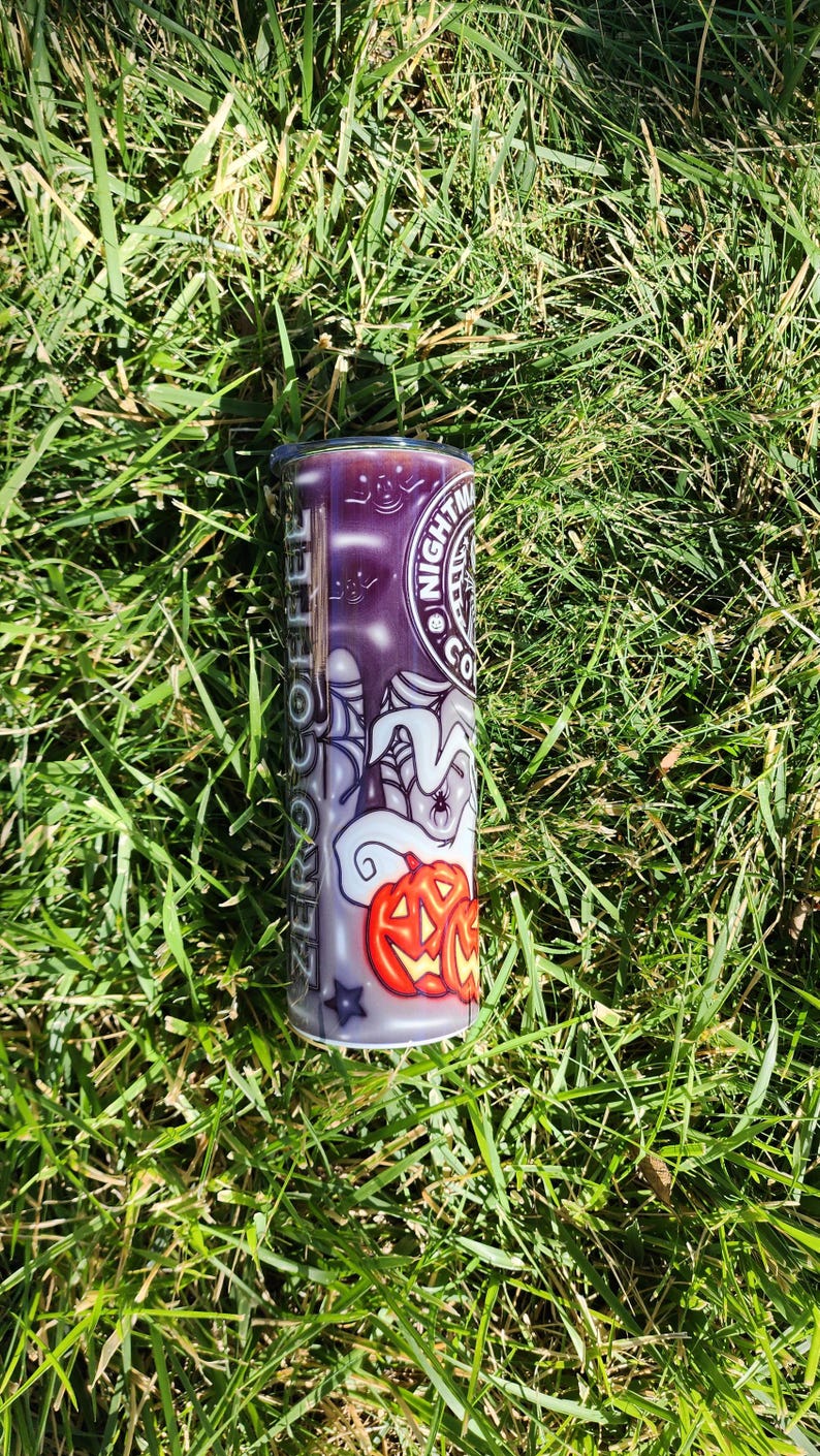 May include: A white and purple tumbler with a Halloween-themed design. The design features a ghost, a spider web, and a jack-o'-lantern. The text "Nightmare Coffee" is printed on the tumbler.