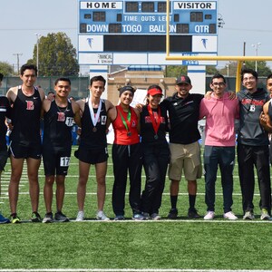 May include: A group of young athletes stand together on a green field, holding medals. They are wearing black and red athletic uniforms with the letters "IB" on their shirts. The athletes are smiling and looking at the camera. The scoreboard behind them reads "HOME" and "VISITOR".