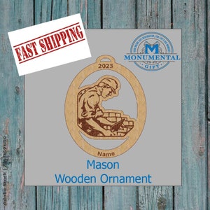 May include: A wooden ornament shaped like a mason laying bricks. The ornament is brown and has a white background. The text "2023", "Name", "Mason", and "Wooden Ornament" is printed on the ornament.