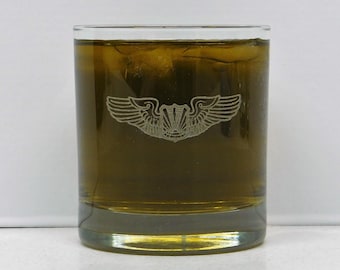 May include: A clear, short glass filled with a golden-brown liquid and ice cubes. The glass features a detailed etched design of a winged emblem. The drink appears to be an alcoholic beverage, possibly whiskey or a similar spirit.