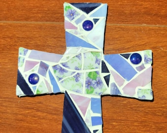Peut inclure: Une croix en mosaïque composée de carreaux bleus, violets et blancs. La croix intègre des carreaux à motifs floraux et des ornements ronds bleu foncé. La mosaïque est posée sur un fond en bois.