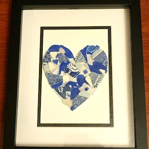 May include: Framed artwork featuring a heart mosaic. The heart is composed of blue and white ceramic shards, set against a white background. The artwork is encased in a black frame, creating a striking contrast.