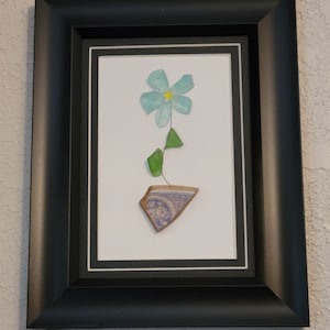 Könnte beinhalten: Gerahmtes Kunstwerk mit einer Blume aus Meeresglas. Die Blume hat eine hellblaue Blüte mit gelbem Zentrum, grüne Blätter und einen Stiel. Die Vase ist ein Fragment aus Keramik mit blauem und violettem Design. Der Rahmen ist schwarz.