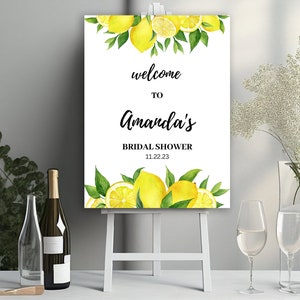 May include: A white easel displays a welcome sign for Amanda's bridal shower, decorated with watercolour lemons and greenery. The sign includes the date 11.22.23. Wine bottles, glasses, and floral arrangements are in the foreground.