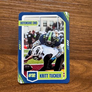 May include: A football trading card featuring a defensive play. The card has a blue border with green and yellow accents. The text "DEFENSIVE END" is at the top, and "#32 KRITT TUCKER" is at the bottom. The image shows two players in action.