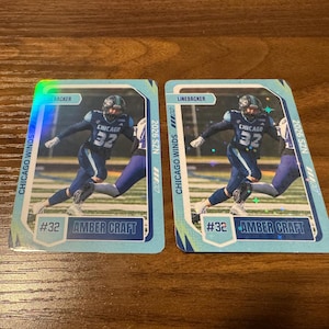 May include: Two identical rectangular sports trading cards featuring a football player in a dark blue uniform with the number 32. The cards display the text Chicago Winds, Linebacker, 2026 SZN, and Amber Craft, with a holographic finish and light blue borders.