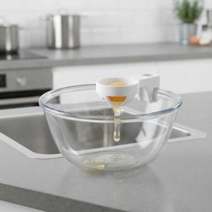 May include: A white plastic egg separator tool is clipped onto the rim of a clear glass mixing bowl. The separator holds a yellow egg yolk while the clear egg white drips through the bottom opening into the centre of the bowl below.