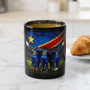May include: A black ceramic mug featuring a graphic of a soccer team in blue jerseys huddled together on a field. The background displays the flag of the Democratic Republic of the Congo and a stadium crowd. The text 2026 is printed at the bottom.