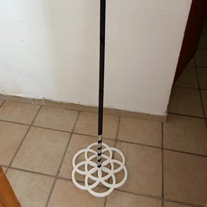 May include: A tall, thin black pole stands vertically, supported by a white, circular base featuring a geometric pattern of seven interlocking rings. The pole has a textured surface and a silver-colored band near the bottom where it meets the base.