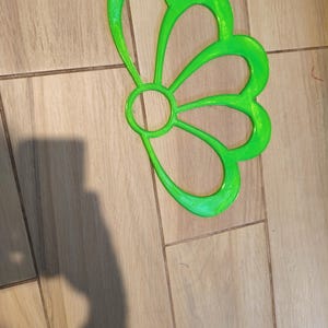 Puede incluir: Una decoración en forma de flor calada, de color verde brillante. El diseño presenta un círculo central con seis formas de pétalos curvados. El objeto está hecho de un material brillante y está sobre una superficie de baldosas marrón claro.