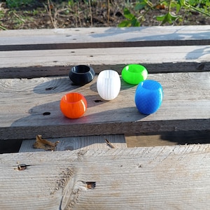 Pode incluir: Uma coleção de objetos coloridos em forma de anel em laranja, preto, branco, verde e azul. Os objetos estão dispostos sobre uma superfície de madeira desgastada, com a luz solar projetando sombras. Os itens parecem decorativos ou funcionais.