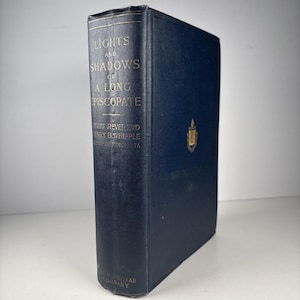 Puede incluir: Un libro de tapa dura antiguo titulado "Lights and Shadows of a Long Episcopate" de Henry B. Whipple. El libro tiene una cubierta azul oscuro con letras doradas y un emblema dorado. El logotipo de Macmillan Company está en la parte inferior.