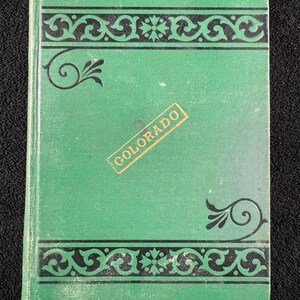 Puede incluir: Un libro verde vintage con bordes y detalles decorativos negros. La palabra "COLORADO" está impresa en una etiqueta rectangular dorada. La cubierta del libro está ligeramente desgastada, lo que sugiere edad y uso.