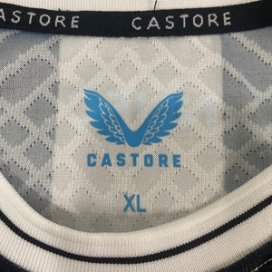 May include: A white and black soccer jersey with a black and white striped pattern on the sleeves. The jersey has a black collar with the text "CASTORE" in white. The front of the jersey features a blue logo with wings and the text "CASTORE" in white. The jersey has a blue tag with the text "XL" in white.
