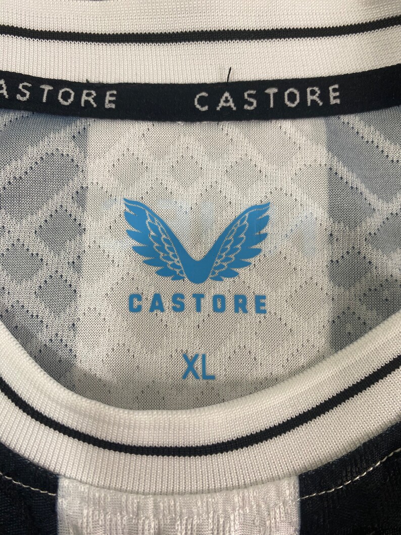 May include: A white and black soccer jersey with a black and white striped pattern on the sleeves. The jersey has a black collar with the text "CASTORE" in white. The front of the jersey features a blue logo with wings and the text "CASTORE" in white. The jersey has a blue tag with the text "XL" in white.