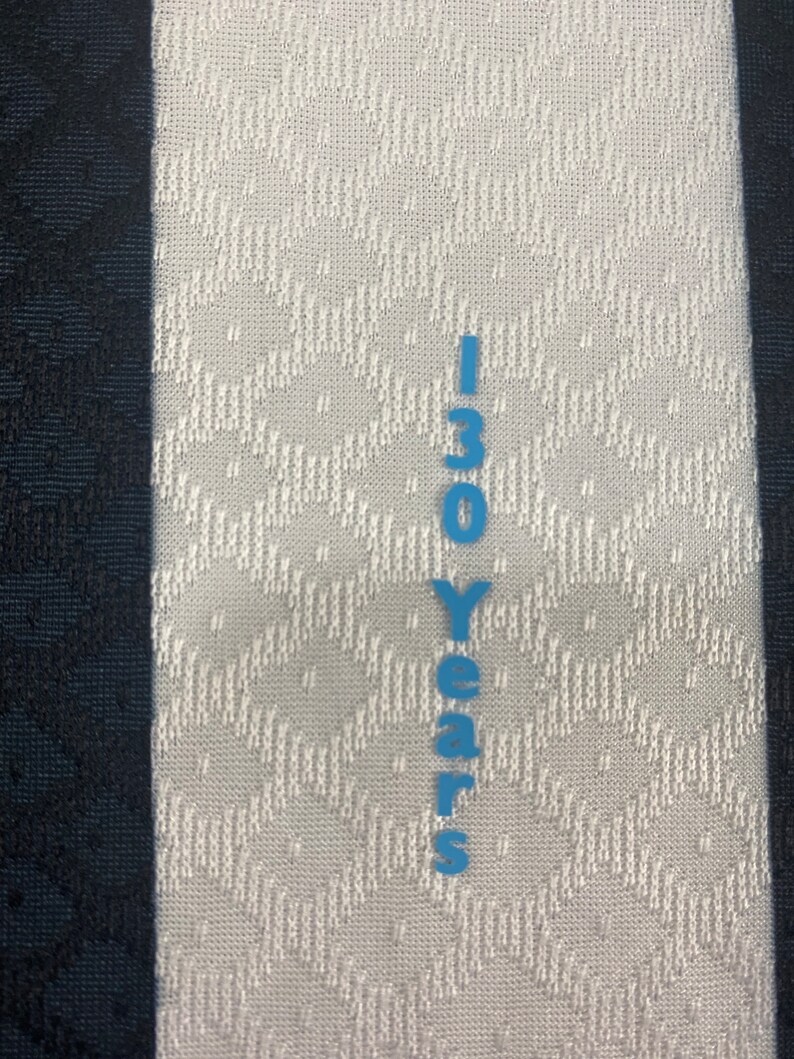 May include: A white fabric with a black stripe on the left side and a blue vertical stripe on the right side. The blue stripe has the text "130 Years" printed on it.