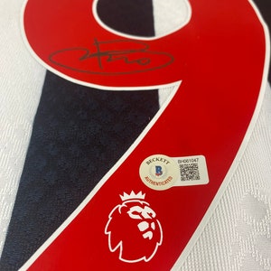 May include: A red and white soccer jersey with the number 9 on the back. The jersey has a black and white striped pattern. The jersey is signed with a black signature. A Beckett Authentication sticker is on the jersey with the text "Beckett Authenticated" and a barcode. The jersey also has a red and white lion crest.