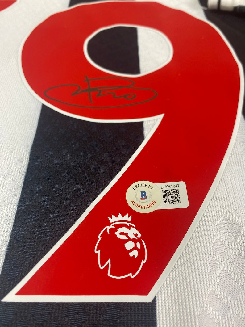 May include: A red and white soccer jersey with the number 9 on the back. The jersey has a black and white striped pattern. The jersey is signed with a black signature. A Beckett Authentication sticker is on the jersey with the text "Beckett Authenticated" and a barcode. The jersey also has a red and white lion crest.