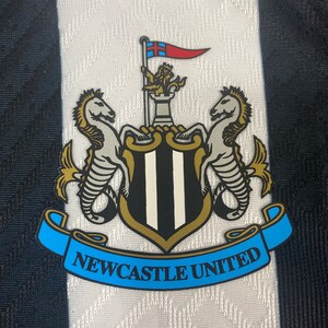 May include: A black and white crest with a gold trim. The crest features two white seahorses flanking a shield with black and white stripes. A gold crown sits atop the shield, and a red and blue flag with a white cross flies above the crown. A blue ribbon with the text "NEWCASTLE UNITED" is wrapped around the bottom of the crest.