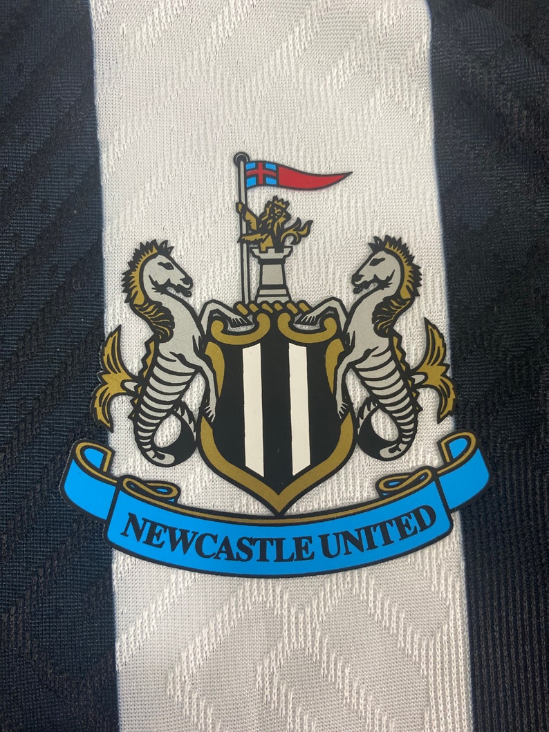 May include: A black and white crest with a gold trim. The crest features two white seahorses flanking a shield with black and white stripes. A gold crown sits atop the shield, and a red and blue flag with a white cross flies above the crown. A blue ribbon with the text "NEWCASTLE UNITED" is wrapped around the bottom of the crest.