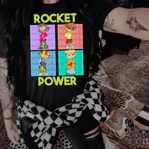 May include: A black t-shirt with a graphic of four cartoon characters in colorful squares. The text "ROCKET POWER" is printed in yellow on the shirt.