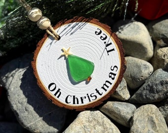 May include: A wooden Christmas tree ornament with the text "Oh Christmas Tree" and a green sea glass Christmas tree. The ornament is sitting on a bed of rocks.